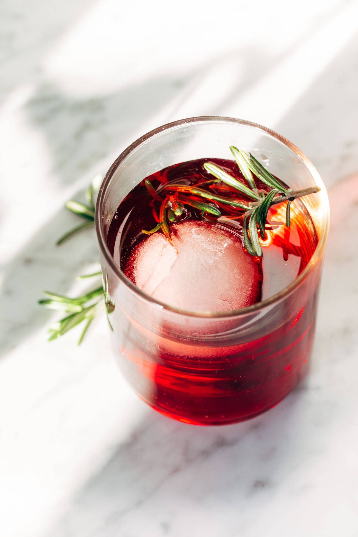 A clear cocktail glass with red liquid, a large ice cube, and a sprig of rosemary.