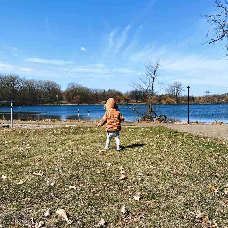 A young child in a brown jacket with the hood over their head walks near the water alone.