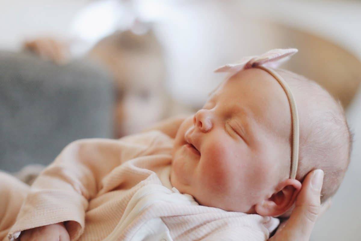 A baby in the hands of Lindsay with her other child in the background.